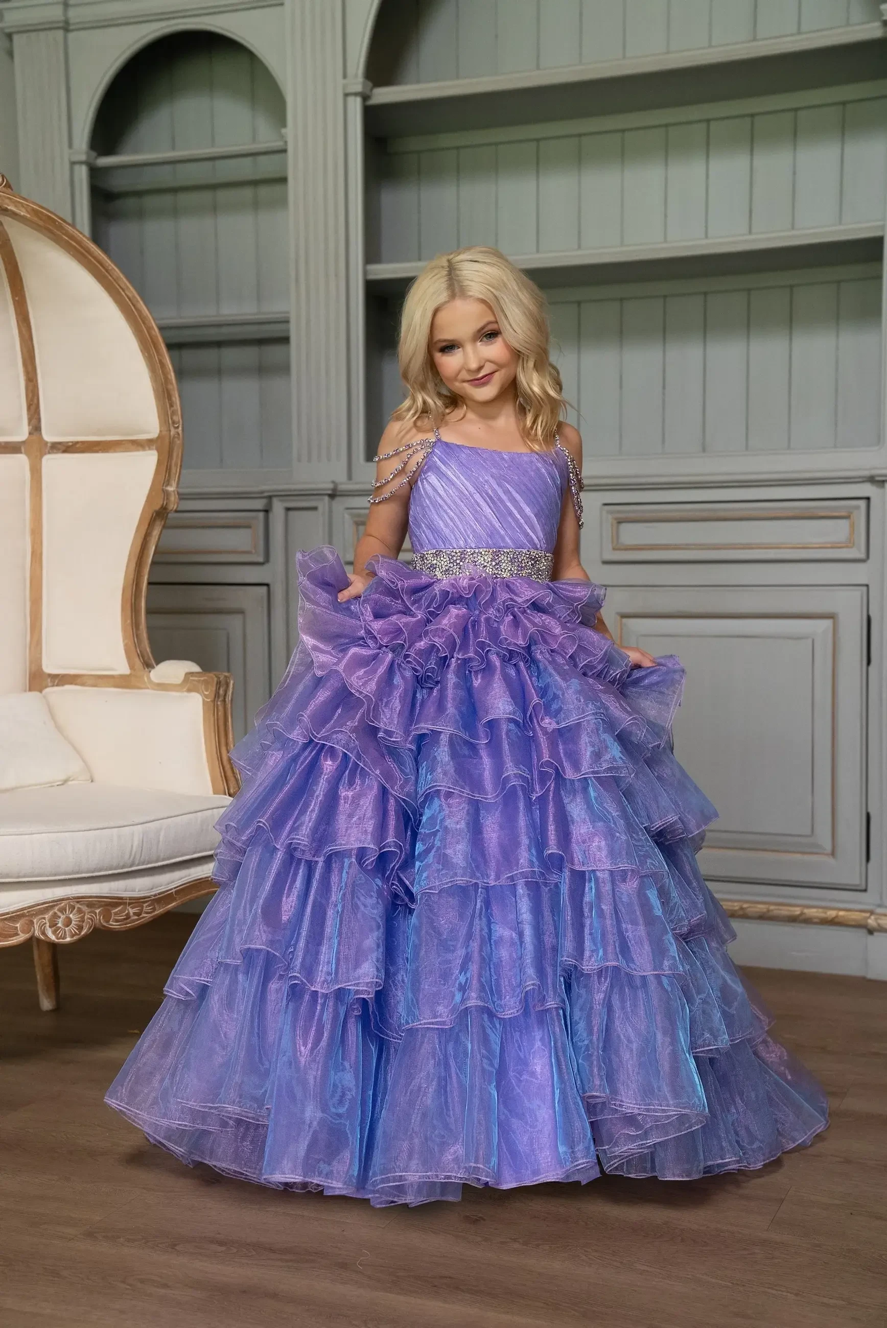 A young girl in a layered, ruffled purple gown stands in a room with decorative furniture.