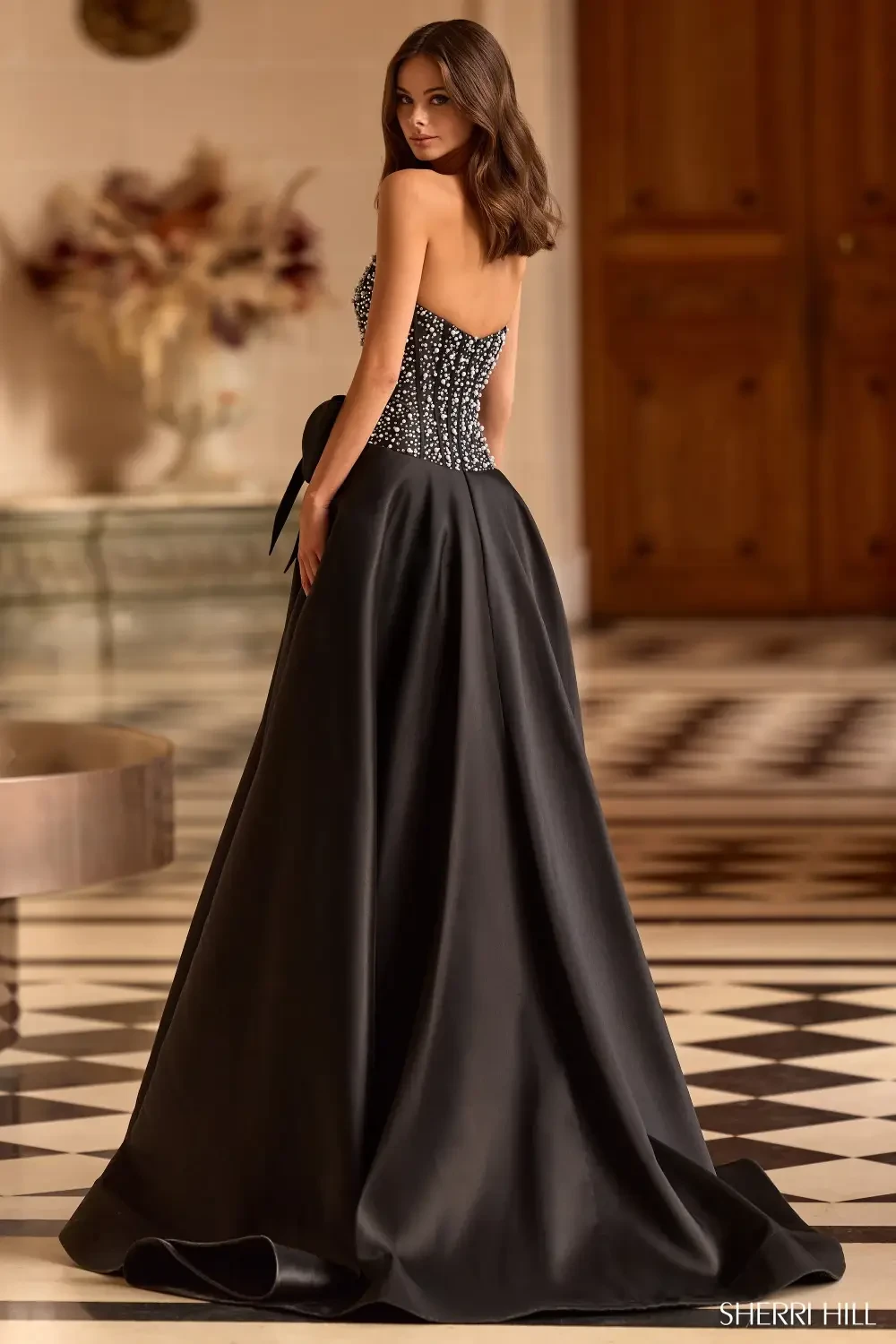 A model wearing a black evening gown with a fitted bodice and a full, flowing skirt, standing with her back to the camera.