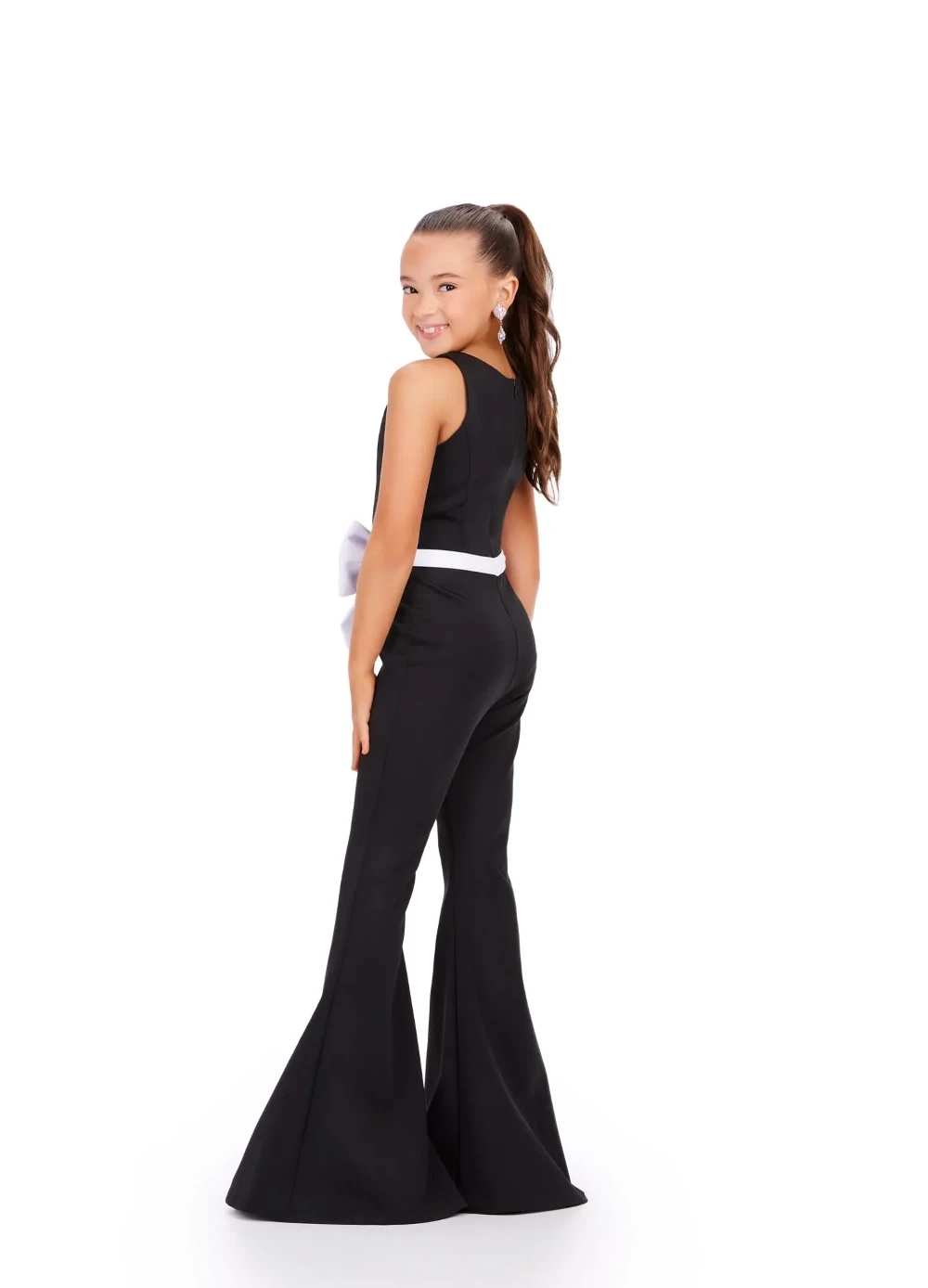A young girl modeling the back of a black pageant interview jumpsuit with flared legs and a white bow detail at the waist.