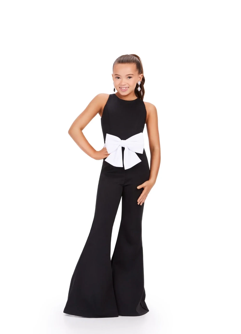 A young girl wearing a black sleeveless pageant interview jumpsuit with flared legs and an oversized white bow at the waist, posing with one hand on her hip.