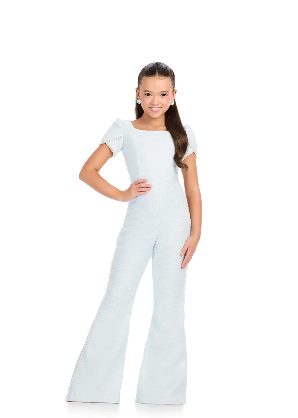 A young girl wearing a light blue pageant interview jumpsuit with short puff sleeves, pearl trim, and flared legs, posing with one hand on her hip.